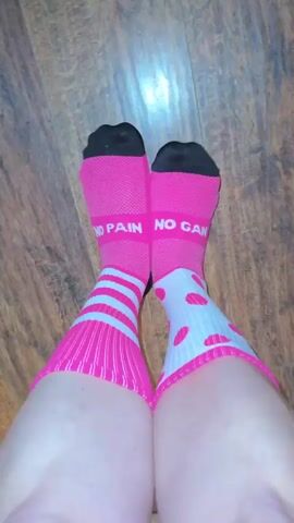 The video shows a close-up, top-down view of a person's legs and feet, which are adorned with bright pink socks. The socks have black toes and heels. The socks on the left leg have horizontal white stripes. The socks on the right leg have white polka dots. Both socks have the words "NO PAIN" in white lettering printed on the calf area. The person's legs are bare above the socks. The background appears to be a wooden floor. The camera is stationary.
