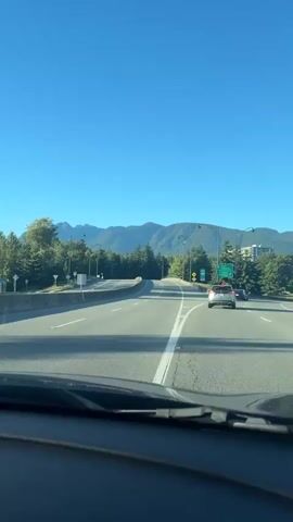 The video shows a view from inside a car driving on a highway. The road ahead is paved with lane markings. There are trees and some buildings on either side of the highway, and mountains in the distance. The sky is clear and blue. There are streetlights lining the highway. A silver car is visible in the right lane ahead of the camera.