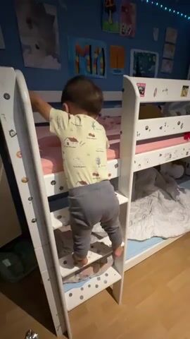 The video shows a toddler climbing a ladder to a bunk bed. The toddler is facing away from the camera, wearing a yellow onesie with a printed design and gray pants. The toddler's left foot is on a ladder rung, and their right foot is on the rung below. The toddler is holding onto the side of the ladder with their left hand and the bunk bed frame with their right hand. The bunk bed has a white frame and pink bedding on the top bunk. The room is decorated with colorful drawings and posters on the blue wall. The floor is wooden. The lighting is dim, with some string lights visible at the top of the wall.