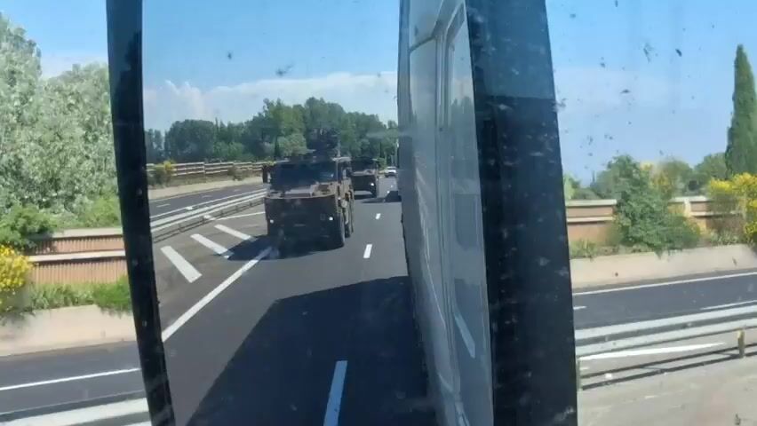 The video shows a side mirror view from a vehicle traveling on a highway. The main focus is the reflection in the mirror, where two military vehicles can be seen moving in the same direction. These vehicles appear to be troop carriers or armored vehicles, painted in a camouflage color. They are driving in the lane to the left of the vehicle from which the video is being recorded. The highway is well-maintained with clear lane markings. The surrounding environment includes green vegetation, trees, and a blue sky with some clouds. Other vehicles can be seen in the background and on the right side of the mirror, including a white truck and a dark SUV. The highway has safety barriers on both sides. The overall scene depicts military vehicles being transported or moving along a public highway during the daytime.
