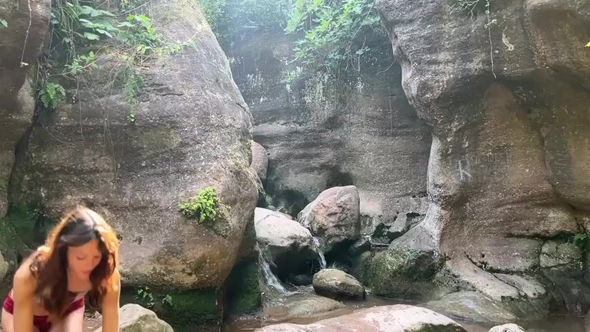 A young woman wearing a red bikini is crouched in a shallow stream at the bottom of a narrow canyon. She is looking down, possibly at the water or something in it. The canyon walls are made of large, rough rocks, with some vegetation growing on them. Sunlight filters down from above, illuminating the scene. There is a small waterfall in the background, flowing over rocks.
