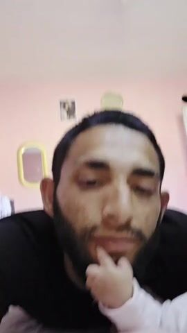 The video features a close-up of an adult male interacting playfully with an infant. The man, with dark hair, a short beard, and a visible neck tattoo on the left side, wears a black t-shirt and an earring in his left ear. His expressions change throughout the clip, starting with a thoughtful gaze, then a slight smile, and concluding with a scrunched-up, playful face with closed eyes, responding to the baby's sounds.

Only a small part of the baby is visible: a hand in a white long-sleeved top resting on the man's chin, and a portion of the baby's head on the right side of the frame. The baby makes various cooing, gurgling, and excited vocalizations throughout the interaction.

The background shows a light pink wall. Decor includes a small, gold-framed oval mirror and a rectangular picture frame with an unclear image hanging on the wall. A ceiling light fixture is visible at the top of the frame. The setting appears to be an indoor, domestic environment. The overall mood is affectionate and playful. No visible text is present.