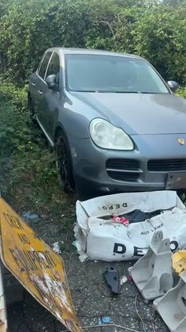 The video shows an outdoor junkyard or salvage yard with several vehicles. The first shot focuses on a gray Porsche Cayenne parked among overgrown weeds. In front of the Porsche is a damaged white cardboard box with the DEPO logo visible. The camera then pans to the right, revealing a dark-colored car with its hood open, partially obscured by foliage. Further to the right, a stack of tires is visible against a chain-link fence. The camera then shifts to a bright red sports car, possibly a Toyota GT86 or Subaru BRZ, with its airbags deployed, indicating it has been in an accident. The red car is parked next to a black SUV. The overall scene suggests a collection of damaged or discarded vehicles in a neglected outdoor environment.