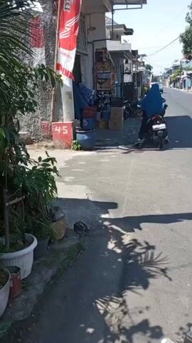 A person wearing a blue hijab is sitting on a black motorcycle parked on the side of a paved road. The motorcycle is facing towards the right of the frame. In front of the motorcycle, there is a small storefront with a red and white banner hanging from it. The storefront has several items displayed outside, including boxes and what appears to be merchandise. To the left of the storefront and motorcycle, there are potted plants and a concrete walkway. The road stretches into the distance, lined with houses and trees. Overhead, there are numerous electrical wires. The sun is shining, casting shadows on the road and ground. The scene appears to be a residential or commercial street in a sunny climate.