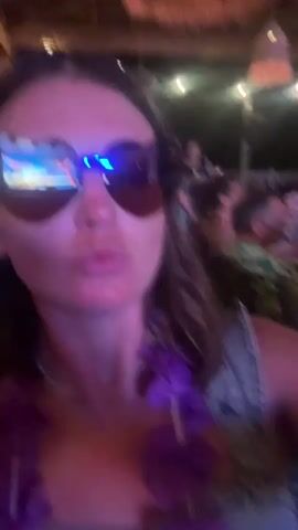 The video shows a woman wearing large heart-shaped sunglasses and a purple lei around her neck. She is in an outdoor setting at night, possibly a luau or similar event, with other people visible in the background. The woman is looking at the camera and making faces, including puckering her lips and making a peace sign. Lights are visible in the background, and there appears to be some sort of thatched roof structure. The overall atmosphere seems festive.