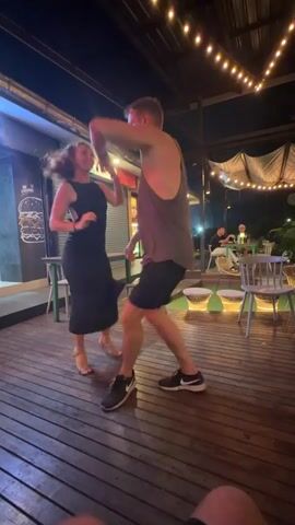 This is a video of a man and a woman dancing on a wooden deck at night. The man is wearing a gray tank top and black shorts, and the woman is wearing a black dress. There are string lights overhead, and a building with a sign that says "BURGER" is visible in the background. The couple appears to be having fun and are smiling at each other.