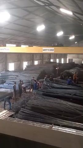 The video shows an interior view of a large warehouse filled with stacks of metal rebar. A yellow overhead crane with the words "SAFETY FIRST" printed on it is visible across the upper section of the warehouse. Several individuals, appearing to be workers, are scattered throughout the space, interacting with the rebar and engaged in various activities. One group of men is clustered together in the center-left, seemingly discussing or examining the rebar. Other individuals are visible in smaller groups or working alone in different areas of the warehouse. The lighting appears to be artificial, coming from overhead fixtures. The overall impression is of an active industrial or construction supply facility.