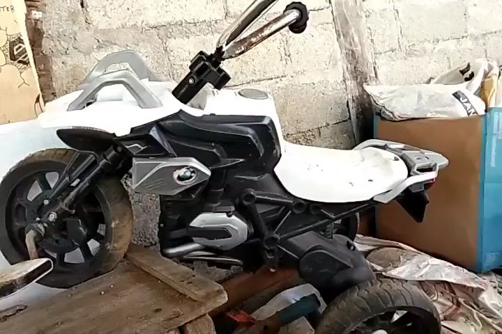 A white toy motorcycle is positioned on wooden planks. The motorcycle has black and silver accents and appears to be a replica of a BMW motorcycle. The background consists of a rough, light-colored wall and various objects, including cardboard boxes and what appears to be a blue tarp. The scene is dimly lit, suggesting an indoor or covered outdoor location.