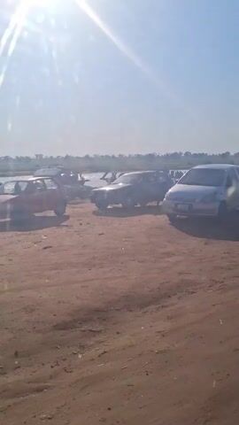 The video shows a line of parked cars on a brown, sandy area near a body of water under a bright, sunny sky. The cars are a mix of colors and models, and the sun is reflecting brightly off their surfaces. In the distance, there appears to be a gathering of people along the shore of the water. The land is mostly barren with sparse patches of vegetation.