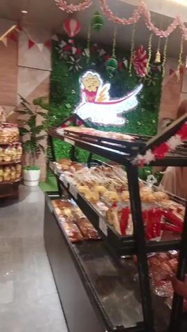 The video shows the interior of a bakery. There are display cases filled with various baked goods. Decorative banners and flags, red and white in color, are displayed throughout the store. People are visible in the background, browsing the merchandise. One person is bending down in the foreground. The lighting is bright and appears to be artificial.