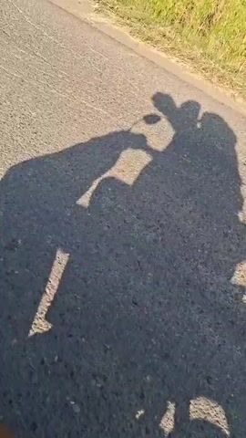 Here is a description of the video:

The video shows the shadow of a person riding a motorcycle down a road. Another person is riding as a passenger. The road is asphalt and lined with tall grass along the side. The shadow moves down the road as the motorcycle drives.
