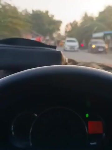 The video shows a driver's perspective from inside a moving car. The dashboard is visible, featuring a speedometer, fuel gauge, and a digital display showing the car's mileage. Through the windshield, a street scene unfolds with other vehicles, motorcycles, and pedestrians moving along the road. The environment suggests an urban or suburban setting, with buildings and trees lining the street. The car appears to be moving at a moderate speed, based on the speedometer reading. A song plays in the background.