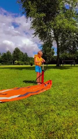 A young boy wearing a yellow life vest and blue shorts stands on a grassy lawn. He is holding a red manual air pump and inflating an orange and black inflatable board, likely a paddleboard or windsurfing board, which lies on the grass. The board is long and flat. In the background, there are green trees and a bright blue sky with white clouds. The sun is shining.