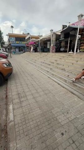 The video shows a street scene with a supermarket in the background. Two women are in the foreground. One woman, wearing a black bra and white shorts, is sitting on the steps leading up to the supermarket. The other woman, wearing a white crochet top and denim shorts, is standing on the steps, facing the supermarket. A white shopping bag is on the steps near the seated woman. Further up the steps, to the right, a man is sitting. The sky is overcast, and palm trees are visible in the background.