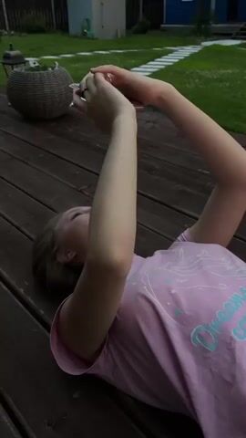 A person wearing a pink t-shirt lies on a wooden deck. The person's arms are raised above their head, and their body is blurred from motion. The background shows a green lawn, a wooden fence, and trees. The sky is cloudy with a vibrant orange sunset visible in the distance. There is a house and a tall structure with a blue dome visible behind the fence.