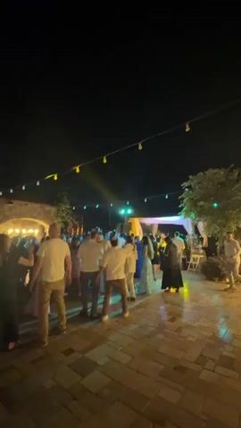 The video begins with a wide shot of an outdoor nighttime gathering, likely a party or wedding reception. String lights are strung overhead, casting a soft glow on a crowd of people dancing and socializing. The ground is paved with light-colored stones. The camera then pans to show a wooden barrel in the foreground, with more people and structures blurred in the background. The perspective shifts to a close-up of a young man with short, light-colored hair, wearing a light-colored button-down shirt. He is looking off-camera with a surprised expression. The video ends with the man speaking, his mouth open, and the camera continuing to focus on his face.