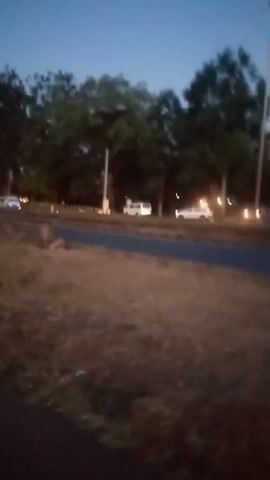 The video depicts a blurry roadside scene at dusk or early evening. A road with a narrow median strip runs through the foreground, with dry, brown grass or dirt in the immediate foreground. Trees and other vegetation can be seen along the roadside, creating a slightly wooded or suburban feel.  Car headlights streak across the road, indicating moving vehicles. The sky is visible above the trees, showing a fading light, suggesting the time of day. The video is shaky and out of focus, giving a sense of movement or recording from a moving vehicle.  The overall lighting is dim.
