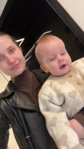 A young woman in a black leather jacket and brown turtleneck holds a baby wearing a cream-colored cardigan with tan hearts. The woman puckers her lips and makes a kissing face, then smiles and speaks. The baby looks directly at the camera and smiles. The background appears to be a white wall with black trim. The video is shot from a close, slightly upward angle.