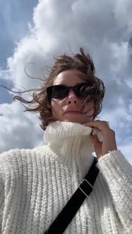 The video shows a medium shot of a young woman from the chest up. She is wearing a white chunky knit turtleneck sweater and black sunglasses. Her brown hair is windswept and styled in a messy bun. She is holding her left hand to her chin, and her right hand is slightly raised. The background is a bright blue sky with large, fluffy white clouds. The woman appears to be outdoors. The video is a selfie-style shot, as the camera is positioned in front of her. The lighting suggests it is daytime.