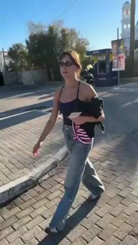 A woman walks on a brick sidewalk on a sunny day. She wears sunglasses, a blue tank top, jeans, and grey shoes. She is carrying a folded black jacket and a striped item. A white building with a blue dome is visible in the background, along with an ATM and parked cars. The sky is clear and blue.