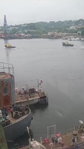 The video shows a harbor with several boats and a crane. In the foreground, there are two docked boats. One is partially visible, with a person standing on its deck. The other boat is more prominent, with a rusty structure and a Russian flag. In the middle ground, two yellow tugboats are visible on the water. One is moving to the left, leaving a wake. The other is stationary or moving slowly to the right. In the background, there is a shoreline with buildings and trees, and a tall crane. The water is calm, and the sky is overcast.