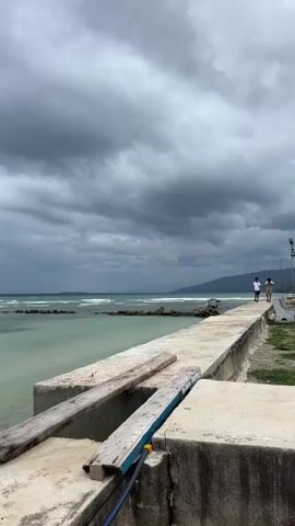 A vertical, slightly shaky shot of a concrete pier extending into the ocean under a cloudy sky. Two people, a man and a woman, walk away from the camera along the pier. The water is a light turquoise color and waves are breaking on a rocky barrier offshore. The pier has some wooden planks resting on concrete blocks in the foreground. The sky is filled with dark, ominous clouds.