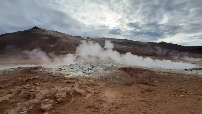The video displays a wide, static shot of an active geothermal field in Reykjahlíð, Iceland, under an overcast sky. The scene is dominated by a barren, reddish-brown landscape, indicative of volcanic activity. Numerous fumaroles and steam vents are scattered across the midground, continuously releasing thick, white plumes of steam and gases that rise into the cool air and drift across the frame.

The ground around these vents features patches of light grey and pale yellow deposits, likely sulfur, contrasting with the dark red-brown earth. In the distance, a range of similarly barren, reddish-brown hills or mountains forms the backdrop under a heavily clouded, grey sky.

A lone individual, small in the vast landscape, can be seen walking from left to right in the mid-distance, partially obscured by the rising steam, providing a sense of scale. In the immediate foreground, the terrain is rugged, rocky, and uneven. A slender red pole or marker is visible sticking out of the ground in the lower-right quadrant of the frame. The time of day appears to be daytime, with diffused light from the overcast conditions. No visible text is present in the video.