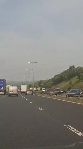 The video shows a highway scene with multiple lanes of traffic. Several trucks and vans are visible, along with some cars. The road appears to be a multi-lane highway with streetlights lining the side. There is a grassy hillside on the right side of the road. The sky is overcast. The vehicles are moving, and it appears to be daytime.