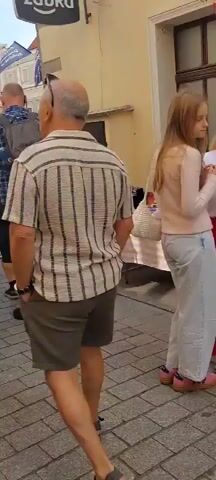 The video shows a street scene that appears to be a market or festival. In the foreground, a young woman with long blonde hair, wearing a light pink long-sleeved shirt and light blue jeans, is looking to her right. Beside her, a woman with blonde hair pulled back, wearing glasses, a white t-shirt, and a long red pleated skirt, is also looking to her right and speaking. Behind them, a man's shoulder and arm are visible on the left side of the frame. In the background, there are stalls with items displayed, tables covered with cloths, and other people walking around. The ground is paved with cobblestones. The buildings in the background are old, with textured walls and windows. There are flags hanging from poles and a traffic light visible. The overall impression is of a busy outdoor event.