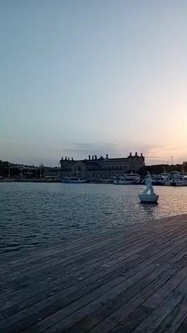 A static, medium shot captures a wide view of a harbor at dusk. The foreground is dominated by a weathered wooden pier, its planks running horizontally across the bottom of the frame. Beyond the pier, a body of calm water stretches to the middle ground, reflecting the soft twilight sky. A white statue of a figure standing on a circular base is visible in the water, slightly to the right of the frame's center. In the background, a long, ornate building with multiple chimneys and arched windows sits along the waterfront, flanked by several boats and smaller buildings. The sky is a gradient of pale blue and soft orange, indicative of the setting sun.