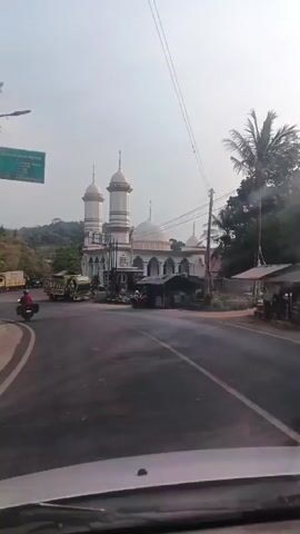 The video shows a mosque with two prominent minarets and a dome. It is situated near a road with traffic, including trucks and motorcycles. The environment appears to be outdoors, with trees and hills in the background. The sky is visible and overcast. The video is likely taken from inside a vehicle, as evidenced by the windshield in the foreground. The overall impression is of a roadside view of a mosque in a potentially rural or semi-urban setting.