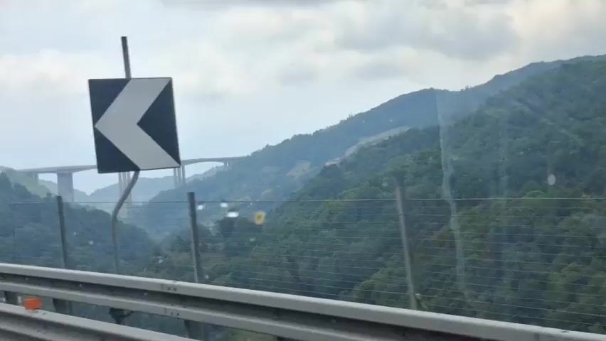 The video shows a view from a vehicle traveling on a highway. In the distance, a large bridge spans across a valley between two forested mountains. A wind turbine is visible on the left side of the frame. The sky is overcast with clouds. The road surface and a wire fence are in the foreground.