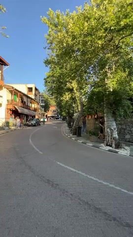 This video captures a serene street scene in Kakopetria, Cyprus, bathed in the warm, golden light of late afternoon or early evening. The sky is a clear, vibrant blue, suggesting pleasant weather. The setting is a charming village street paved with cobblestones, curving gently to the right.

Dominating the foreground are large, mature trees with lush green foliage, their trunks gnarled and textured. Their branches cast dappled shadows across the road and pavement. Along the right side of the street, a low stone wall supports a raised area with a wooden fence and an outdoor seating area, possibly a cafe or restaurant, indicated by umbrellas and wooden structures. A lamppost stands near the stone wall.

Several people are visible, engaged in casual activities. On the left, a few individuals are seen walking along the sidewalk next to buildings that appear to be shops or houses, some with balconies. Further along the street, a person is pushing a stroller, indicating a leisurely stroll. Another individual walks a bit ahead. The overall mood is peaceful and relaxed, evoking a sense of tranquil village life.

There are no visible text elements in the video. The location cues, such as the architecture and the stone walls, along with the context provided, firmly place the scene in Kakopetria, Cyprus. The lighting and the presence of people enjoying the outdoors suggest a warm day with a comfortable evening approaching.