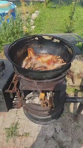 A close-up shot shows pieces of rabbit being fried in a large black cast-iron pot. The pot is heated by a fire burning in a metal drum below. The rabbit pieces are browning and sizzling in hot oil, with some parts already appearing golden brown. The scene is set outdoors with green foliage visible in the background. The fire is producing smoke and flames, indicating active cooking. The overall impression is of a rustic outdoor cooking setup.