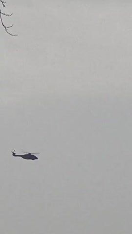 A dark helicopter with its rotors spinning is visible in the sky. A building is partially visible on the right side of the frame. The sky is overcast and appears light gray.