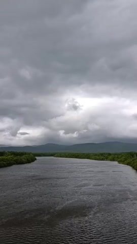 A wide, dark river flows through a landscape of lush green trees and rolling hills under a cloudy sky. The camera is positioned on a bridge overlooking the scene. The water's surface is calm, reflecting the overcast sky. Trees line both banks of the river, creating a dense wall of green that extends to the horizon where mountains are visible in the distance. The clouds are heavy and grey, suggesting an impending storm or a recent rainfall. A red and white railing from the bridge is visible in the foreground on the right side of the frame. The overall atmosphere is serene but somber due to the weather.