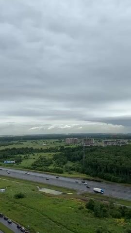 The video is a vertical, slightly shaky aerial shot of a highway and surrounding landscape under a cloudy sky. The highway is in the foreground, with several cars and a white truck driving on it. To the left of the highway is a green grassy area with some small puddles. To the right of the highway is a dense forest. In the middle ground, there are more fields and trees, and in the background, there are several large buildings under construction and a few completed buildings. The sky is overcast with gray clouds, and there appears to be a hint of sunlight breaking through in the distance.