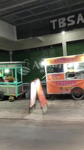 The video captures a bustling street scene in Sumorame, East Java, likely in the evening due to the artificial lighting. The focus is on a row of shops and street vendors.

In the foreground, a motorcycle is seen passing by, indicating vehicular traffic. To the left, a food cart with a light blue and green frame is illuminated, with a person visible inside, possibly preparing or serving food. Further down the street, another food cart with a yellow and blue frame is also present, with a person standing behind it.

The shops themselves have distinctive signage. One shop has a large green facade with white lettering that appears to read "avian BRANDS". Another shop has a yellow awning with the word "PETSHOP" clearly visible in bold lettering. The shop interiors are visible, showing shelves stocked with items, suggesting a retail environment.

The street surface appears wet in some areas, possibly from recent rain or cleaning, reflecting the lights and creating a slightly hazy effect in the video. The overall atmosphere is one of commercial activity, with vendors and shops open for business. The time of day appears to be late afternoon or early evening, as suggested by the warm artificial lighting and the general ambiance.