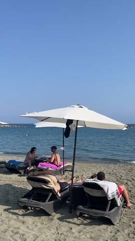The video shows a beach scene on a clear, sunny day. The sky is a deep blue, and the ocean is calm and blue with gentle waves. The beach is sandy and there are several people relaxing. In the foreground, two lounge chairs with white umbrellas are positioned on the sand. A man in a white t-shirt and red shorts is sitting in the right lounge chair, facing away from the camera and towards the ocean. A man in a black t-shirt and shorts is lying in the left lounge chair, also facing away from the camera. Behind these two chairs, a man wearing a dark t-shirt and shorts is sitting on a purple mat. To his left, a woman in a pink top and a white skirt is also sitting on a purple mat. In the distance, a person is visible in the water. A man is on a jet ski or similar watercraft. There are some rocks or a breakwater in the far distance. The overall atmosphere appears relaxed and serene.