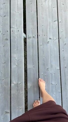 The video shows a person's bare feet and legs as they walk on a wooden deck. The deck planks are grey and weathered, with visible grain and knots. There is a gap between two of the planks, through which some dark ground or vegetation is visible. The person is wearing a dark maroon garment, possibly a hoodie or dress. The camera is positioned above and behind the person, looking down at their feet and the deck. The lighting suggests it is daytime. The person's toenails are painted with white polish.