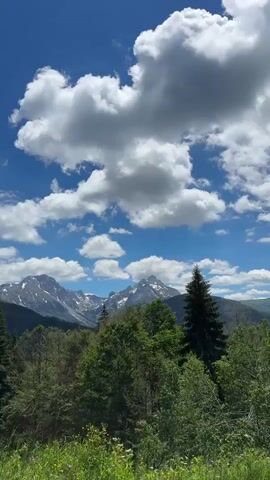 The video shows a majestic mountain landscape under a bright blue sky filled with fluffy white clouds. In the foreground, lush green pine trees are visible, with some deciduous trees interspersed. A rustic, metal fence runs horizontally across the lower part of the frame, suggesting a boundary or enclosure. The background is dominated by imposing, snow-capped mountains with rugged peaks, hinting at their grandeur and altitude. The overall impression is one of natural beauty and tranquility in a mountainous region. The camera appears to be panning slightly, shifting the perspective to reveal more of the expansive scenery.