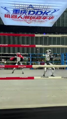Two humanoid robots are sparring in a boxing ring. The robot on the left wears red boxing gloves and a red headguard, while the robot on the right wears white boxing gloves and a white headguard. Both robots are in fighting stances, with their fists raised. The ring is surrounded by ropes, and there is a large screen in the background with Chinese text and a star logo. The environment appears to be indoors, with lights illuminating the scene.