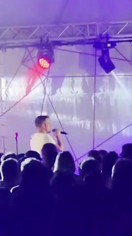 A man is singing into a microphone on a stage at a concert. He is wearing a white t-shirt. The stage is lit with red and white lights, and there is smoke in the air. A crowd of people are visible in the foreground, silhouetted against the stage lights. The man is holding the microphone with his right hand and gesturing with his left. The lighting changes from red to white to purple.