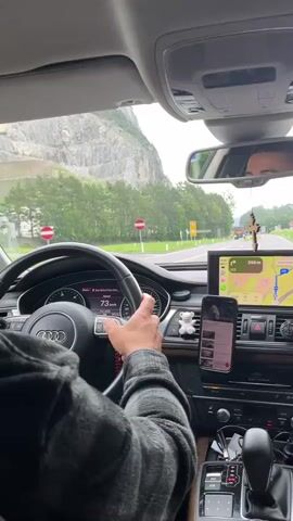 The video shows the interior of a car, looking forward through the windshield. The driver's hands are visible on the steering wheel. The car is moving along a road with trees and a mountainous landscape on the left. There are road signs visible. The dashboard of the car is visible, including a navigation screen displaying a map and a phone mounted to the dashboard. The speed displayed on the dashboard is fluctuating between 73 km/h and 69 km/h.