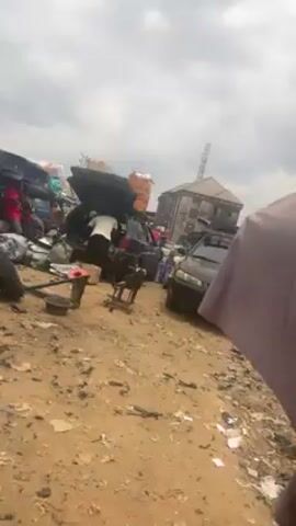 The video shows a busy outdoor marketplace with cars loaded with goods, tires, and other items scattered around. There is a man carrying a large object on his head. A sign advertising financial services is visible. The ground is dirty and littered.