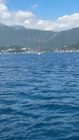 A white sailboat with a prominent red mast is anchored or slowly drifting on a body of blue water. The water has gentle ripples, indicating calm conditions. In the background, lush green mountains rise, with several white and light-colored houses and buildings visible along the coastline at their base. The sky is partly cloudy with patches of blue. The scene depicts a serene daytime setting in a coastal area, specifically Luštica, Montenegro. No people are clearly visible on the boat, and no text is present in the video.