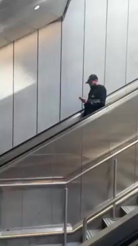 The video shows two men on an escalator in what appears to be a modern building with stainless steel and white paneling. The first man, wearing a black baseball cap, a black long-sleeved shirt, and carrying a beard, is looking at his phone as he ascends the escalator. Behind him, a second man with gray hair and sunglasses, wearing a gray jacket, also rides the escalator. The camera is positioned at a low angle, looking up at the escalator. The lighting is bright, with natural light filtering into the scene.
