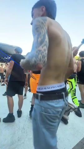 The video shows a group of young adults at an outdoor event, possibly a concert or festival, with a clear blue sky in the background. The focus is on a shirtless young man with tattoos on his arm, wearing denim shorts and white boxer briefs. He is dancing and appears to be holding a drink in his hand. Other individuals are visible in the background, some also dancing and some standing. The environment suggests a daytime event with casual attire and a lively atmosphere.
