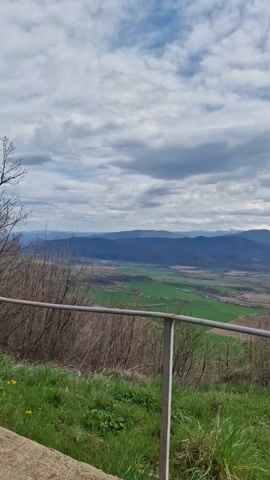 The camera slowly pans right, revealing a verdant valley overlooked by a range of mountains capped with snow. A sky with scattered white clouds provides the backdrop. In the foreground, a metal railing and sparse vegetation frame the view. To the right, a blue observation platform extends out, offering an unobstructed panorama.