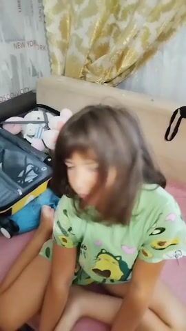 This media file shows a young girl sitting on a bed with an open suitcase beside her. The girl has dark hair and is wearing a light green pajama top with avocado and heart prints. She is looking at the camera with a neutral expression.

The suitcase is partially packed with stuffed animals, including a brown bear, a pink bunny, and a blue dinosaur toy on top of the suitcase. The background shows a wall with a textured wallpaper featuring city names like "DUBAI," "MOSCOW CITY," and "NEW YORK." There are also curtains in the background.

The scene appears to be indoors, likely in a bedroom. The lighting suggests it could be daytime. The presence of the suitcase and packed stuffed animals implies that the girl might be preparing for or has recently returned from a trip. The overall mood of the image is calm and domestic.